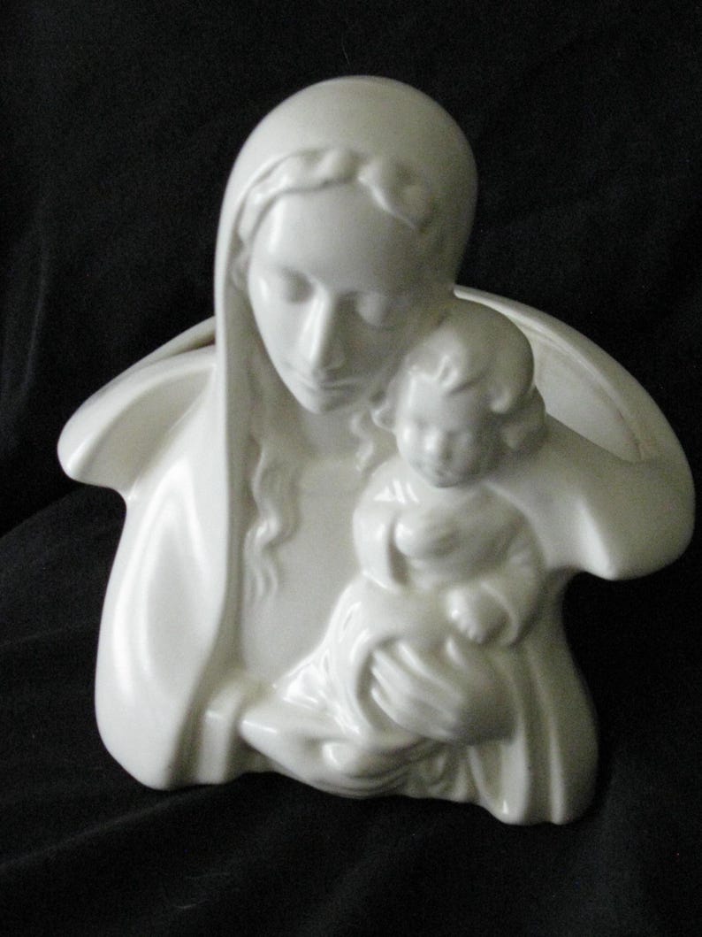 May include: A white ceramic sculpture depicting a mother holding a child. The figures are draped in flowing garments, with the mother's head covered by a veil. The sculpture has a smooth, glossy finish and is set against a dark background.