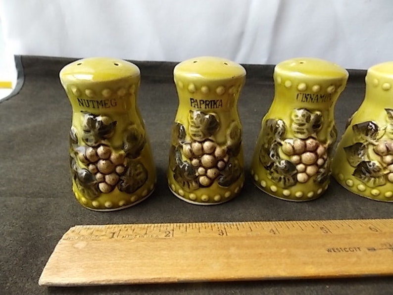May include: Set of four ceramic spice shakers in a light green color. Each shaker is labeled with a spice name: nutmeg, paprika, and cinnamon. The shakers have a grape design and are approximately 3 inches tall.
