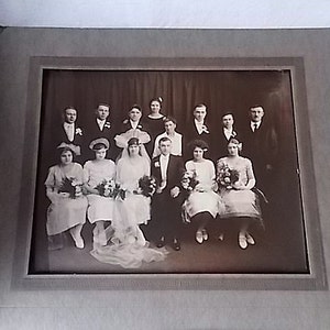May include: A vintage black and white photograph of a wedding party. The bride and groom are seated in the center, surrounded by bridesmaids and groomsmen. The photo is in a gray frame, and the subjects are dressed in formal attire.