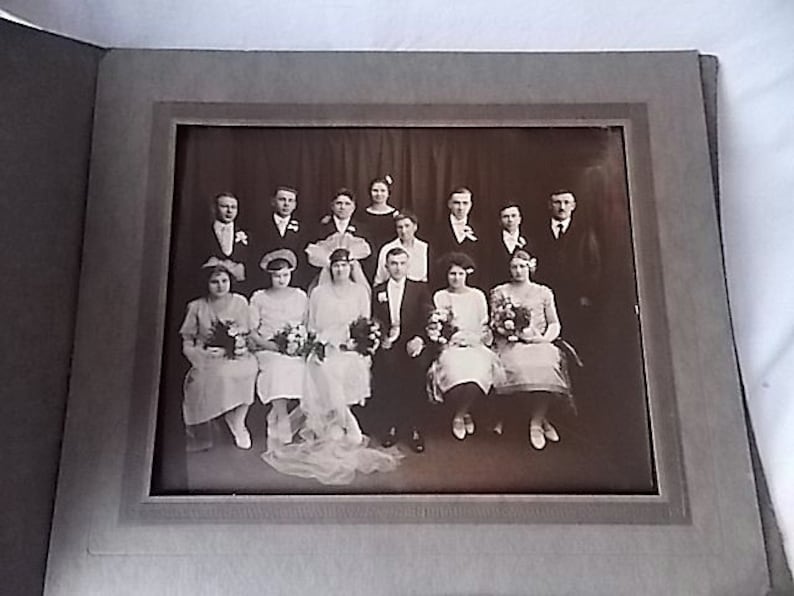 May include: A vintage black and white photograph of a wedding party. The bride and groom are seated in the center, surrounded by bridesmaids and groomsmen. The photo is in a gray frame, and the subjects are dressed in formal attire.