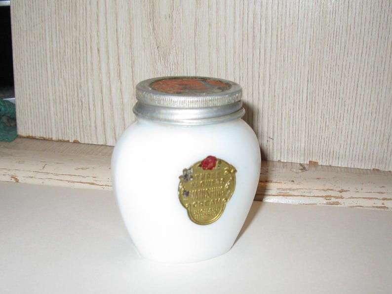 May include: A white, opaque glass jar with a silver lid and a gold label. The label has red and gold accents and text that reads "Sunshine Cream". The jar is likely an antique cosmetic container.