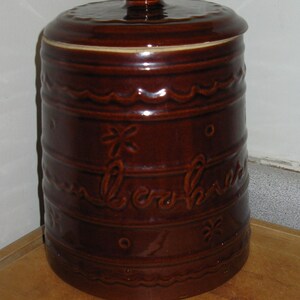 May include: A brown ceramic cookie jar with a lid. The jar has a cylindrical shape with decorative wavy lines, floral accents, and the word "Cookies" embossed on the side. The jar is sitting on a wooden surface.