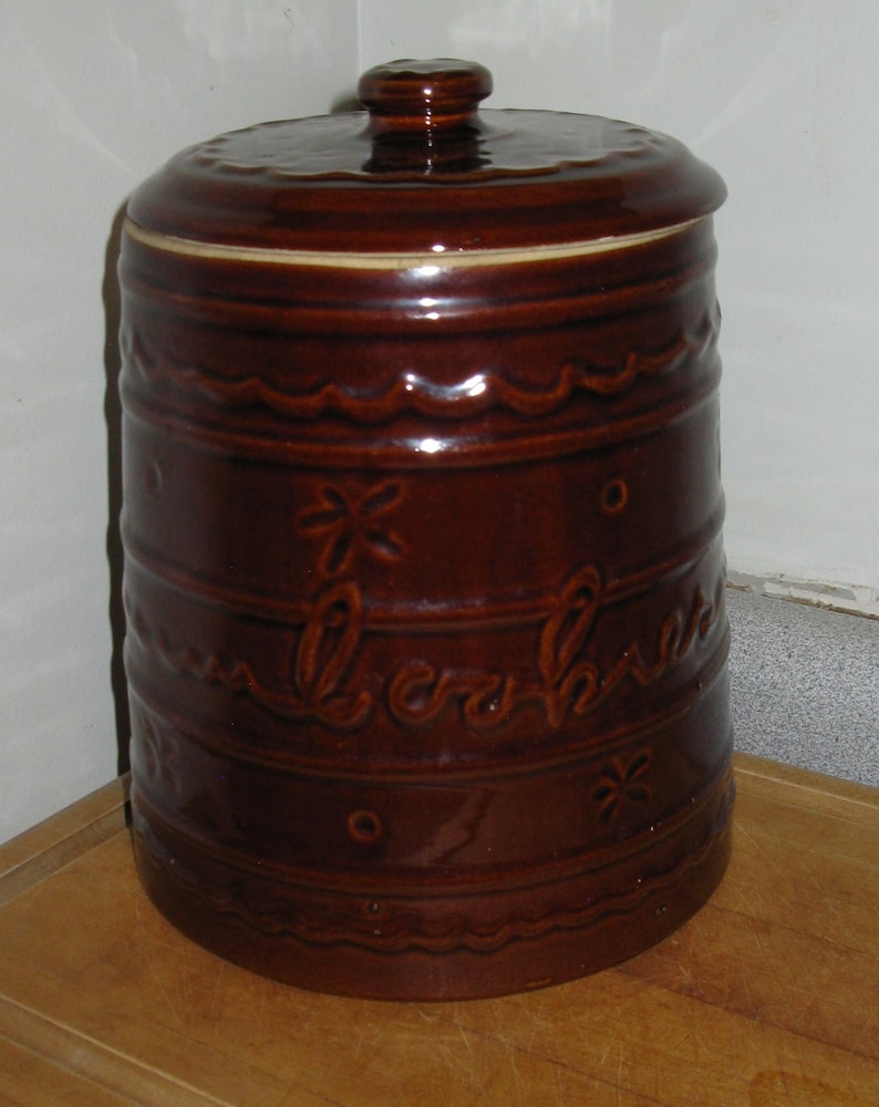 May include: A brown ceramic cookie jar with a lid. The jar has a cylindrical shape with decorative wavy lines, floral accents, and the word "Cookies" embossed on the side. The jar is sitting on a wooden surface.