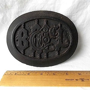 May include: Dark brown, oval metal object with raised lettering and a textured surface. The letters "BCA" are visible in the center. A wooden ruler with inch markings is in the foreground.