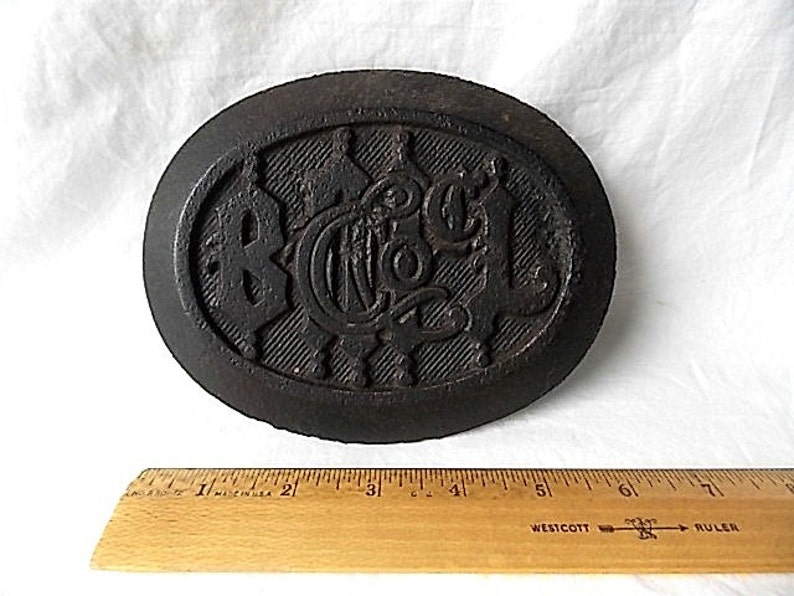 May include: Dark brown, oval metal object with raised lettering and a textured surface. The letters "BCA" are visible in the center. A wooden ruler with inch markings is in the foreground.