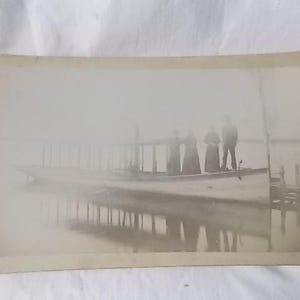 May include: Vintage sepia-toned photograph of a boat with several people on board. The boat is on the water, and the reflection of the boat and the people is visible. The image has a soft, hazy quality, suggesting an older photograph.