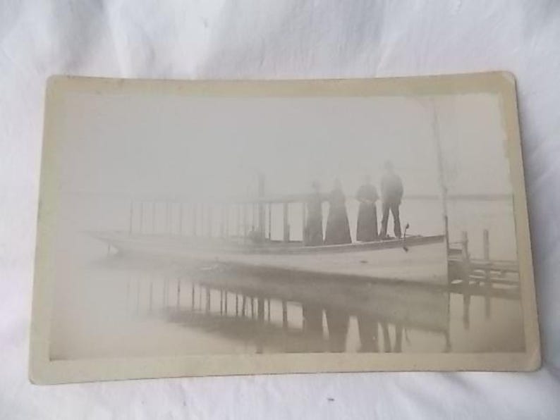 May include: Vintage sepia-toned photograph of a boat with several people on board. The boat is on the water, and the reflection of the boat and the people is visible. The image has a soft, hazy quality, suggesting an older photograph.