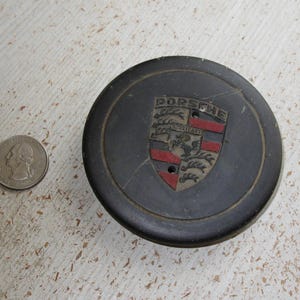 Puede incluir: Un tapacubos negro de Porsche con el emblema de Porsche en el centro, con un escudo con detalles rojos, negros y dorados. Una moneda de veinticinco centavos estadounidense est&aacute; al lado para comparar el tama&ntilde;o.