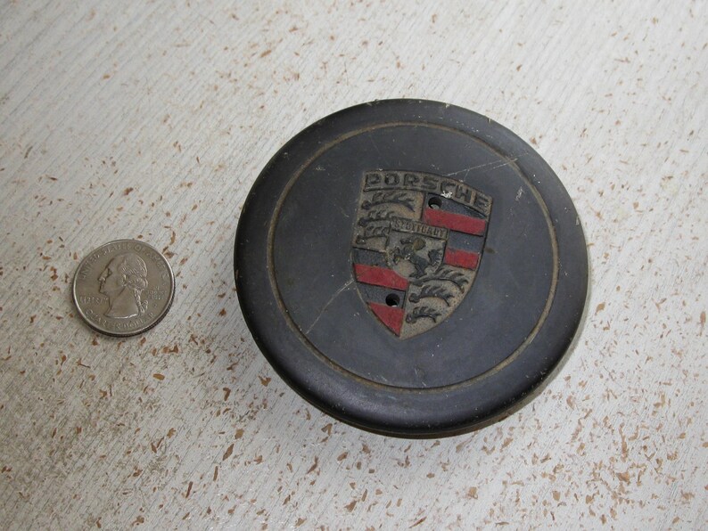 Puede incluir: Un tapacubos negro de Porsche con el emblema de Porsche en el centro, con un escudo con detalles rojos, negros y dorados. Una moneda de veinticinco centavos estadounidense est&aacute; al lado para comparar el tama&ntilde;o.