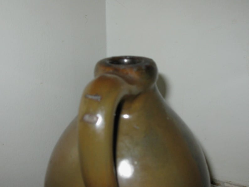 Pode incluir: Um jarro de cer&acirc;mica castanho com uma pega. O jarro tem um corpo arredondado e um gargalo estreito. A pega &eacute; fixada na lateral do jarro e curva-se para cima. O jarro &eacute; feito de um material cer&acirc;mico vidrado.