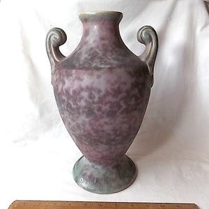 May include: A speckled purple vase with two curved handles. The vase has a wide base and a narrow neck with a light green rim. A wooden ruler is at the bottom of the image. The vase is likely for decorative purposes.