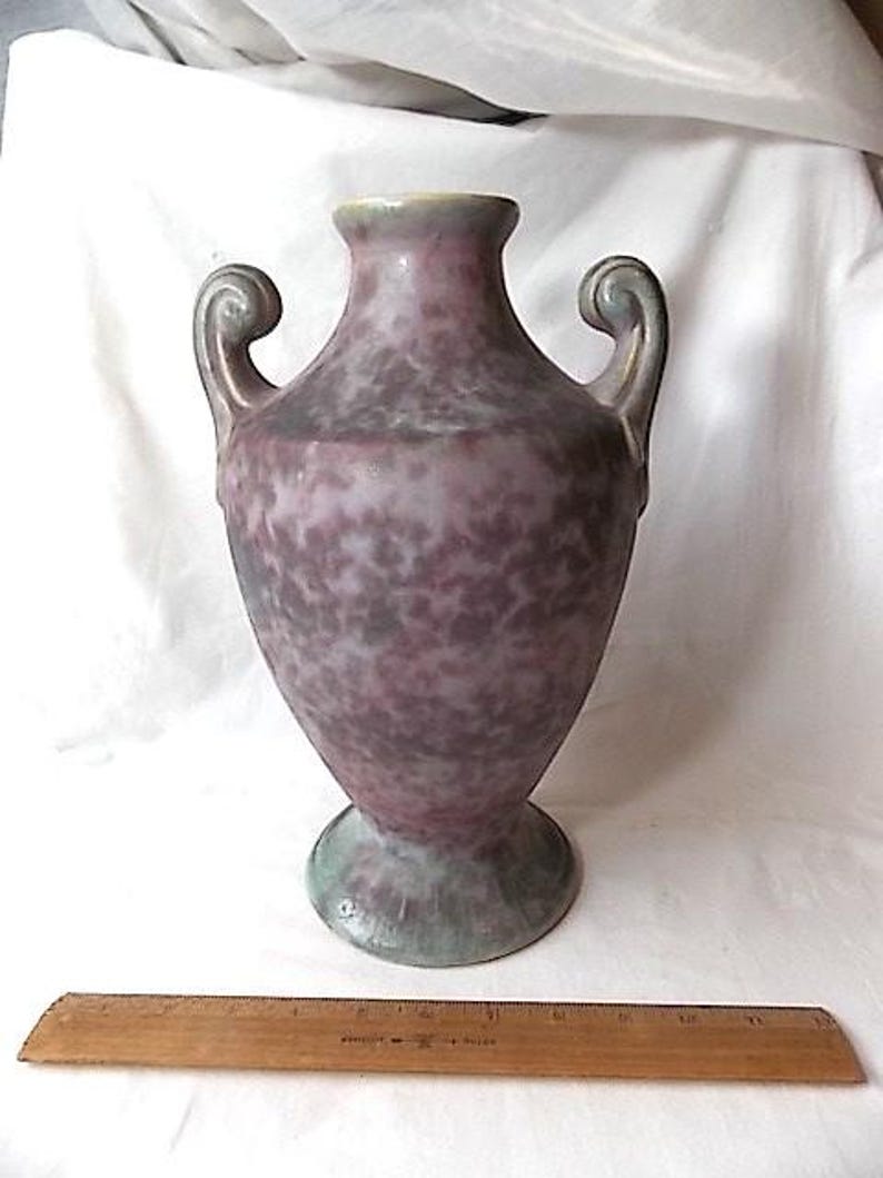 May include: A speckled purple vase with two curved handles. The vase has a wide base and a narrow neck with a light green rim. A wooden ruler is at the bottom of the image. The vase is likely for decorative purposes.