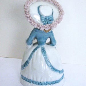 May include: A porcelain figurine of a person wearing a blue jacket and a white skirt with blue trim. The figure wears a wide-brimmed hat featuring a light pink ruffled edge and a blue bow detail. The design includes a flared skirt silhouette.