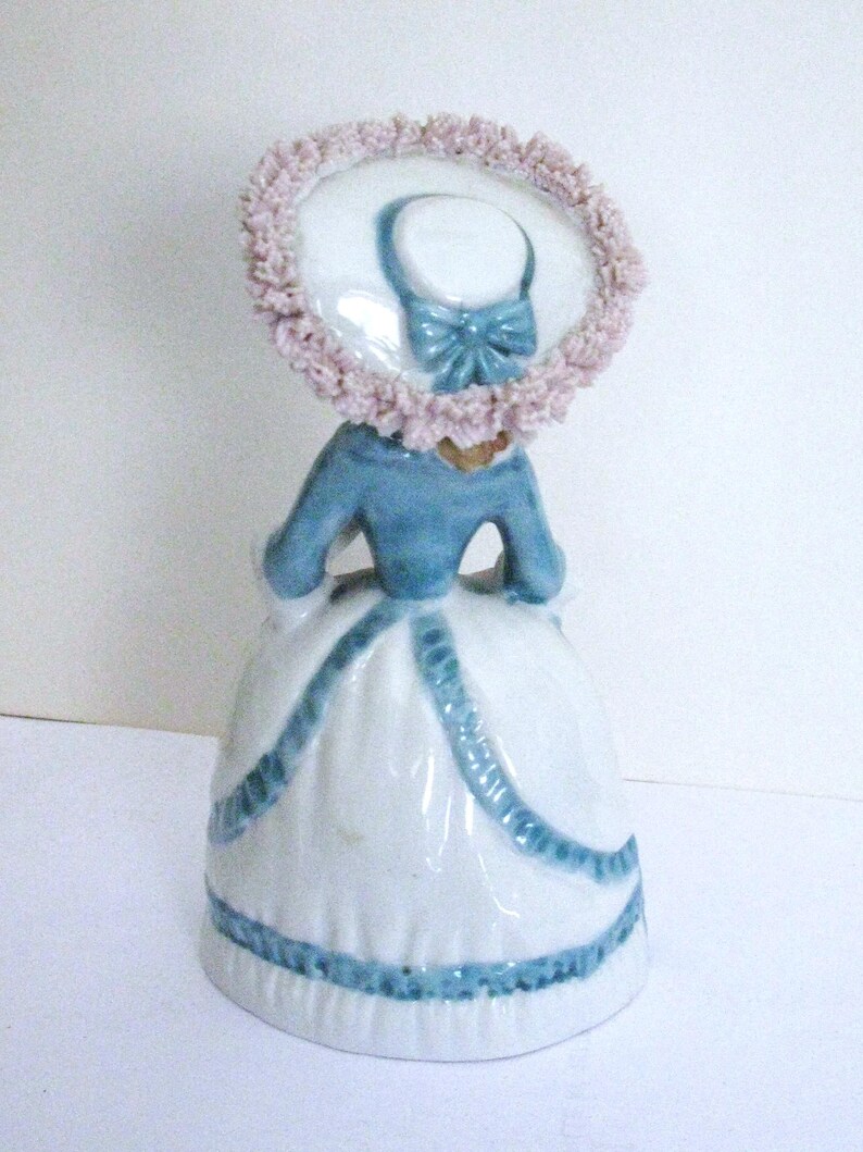 May include: A porcelain figurine of a person wearing a blue jacket and a white skirt with blue trim. The figure wears a wide-brimmed hat featuring a light pink ruffled edge and a blue bow detail. The design includes a flared skirt silhouette.