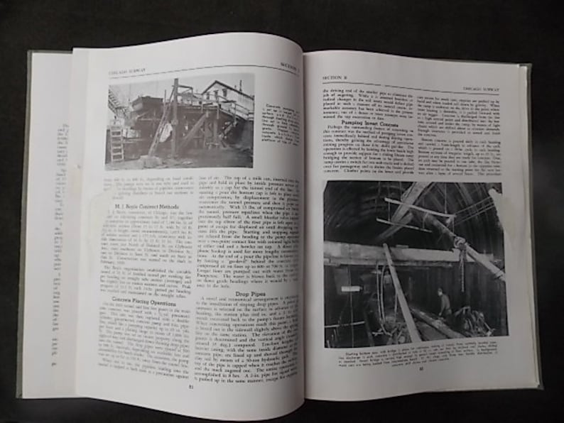 May include: An open book with black and white photographs and text. The left page features a photo of a construction site with a ladder. The right page shows a photo of a wooden structure. The book is open on a dark surface.