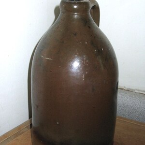 Pode incluir: Um jarro de gr&eacute;s castanho com corpo arredondado e uma pega. O jarro tem um acabamento escuro e brilhante e uma pequena abertura no topo. &Eacute; provavelmente uma pe&ccedil;a antiga ou vintage, adequada para decora&ccedil;&atilde;o r&uacute;stica.