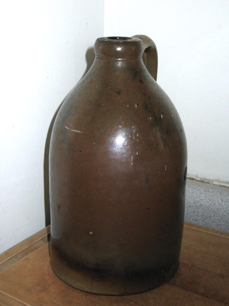 Pode incluir: Um jarro de gr&eacute;s castanho com corpo arredondado e uma pega. O jarro tem um acabamento escuro e brilhante e uma pequena abertura no topo. &Eacute; provavelmente uma pe&ccedil;a antiga ou vintage, adequada para decora&ccedil;&atilde;o r&uacute;stica.