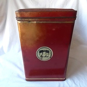 May include: A rectangular red metal tin with a lid, featuring a circular label on the front with the text Aeska and a graphic of a person. A wooden ruler is positioned at the base, showing the tin is approximately 6 inches in height.