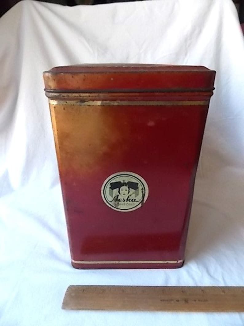 May include: A rectangular red metal tin with a lid, featuring a circular label on the front with the text Aeska and a graphic of a person. A wooden ruler is positioned at the base, showing the tin is approximately 6 inches in height.