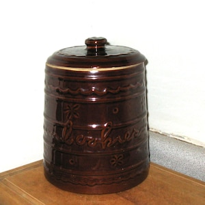 May include: A brown ceramic cookie jar with a lid. The jar has a cylindrical shape with embossed floral and wave patterns. The word "cookies" is written on the side in cursive script. The jar is sitting on a wooden surface.