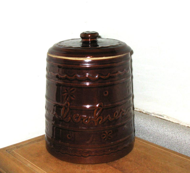 May include: A brown ceramic cookie jar with a lid. The jar has a cylindrical shape with embossed floral and wave patterns. The word "cookies" is written on the side in cursive script. The jar is sitting on a wooden surface.