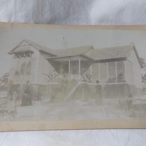May include: Vintage black and white photograph of a house with a porch and several people standing in front. The house has a gabled roof and a screened-in porch area. The photograph is slightly faded.