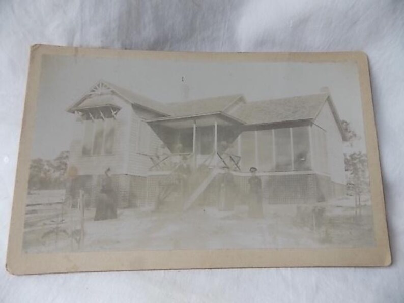 May include: Vintage black and white photograph of a house with a porch and several people standing in front. The house has a gabled roof and a screened-in porch area. The photograph is slightly faded.