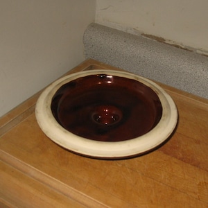 May include: A brown ceramic bowl with a cream-colored rim sits on a wooden surface. The bowl's interior is a deep brown, and it has a central, raised design. The bowl is likely for food or decorative purposes.