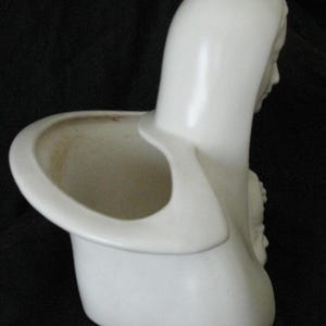 May include: A white ceramic planter in the shape of a woman's bust, featuring a hooded head and a circular opening for plants. The planter has a smooth, glossy finish and a detailed relief on the side.