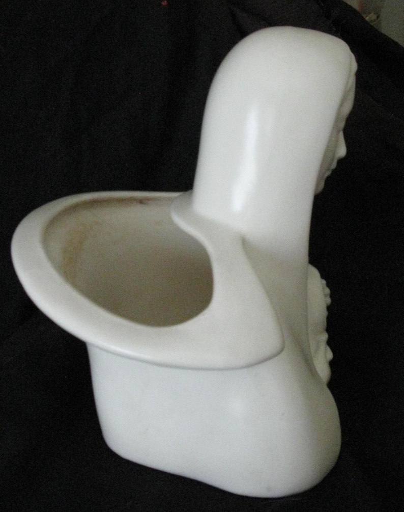 May include: A white ceramic planter in the shape of a woman's bust, featuring a hooded head and a circular opening for plants. The planter has a smooth, glossy finish and a detailed relief on the side.