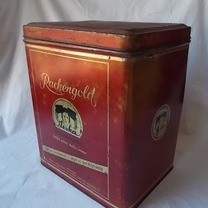 May include: A rectangular red metal tin with a lid, featuring the gold text "Rachengold" and a circular graphic of a person. The container displays additional German text and a smaller circular logo on the side, showing signs of vintage wear.