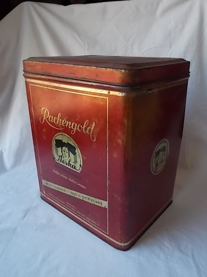 May include: A rectangular red metal tin with a lid, featuring the gold text "Rachengold" and a circular graphic of a person. The container displays additional German text and a smaller circular logo on the side, showing signs of vintage wear.