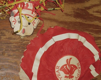 May include: A vintage New Year's party favor set featuring a paper cutout of a face with red and gold fringe hair and a red pleated paper fan with a circular center label depicting two bells and the text Happy New Year in red ink.