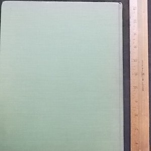 May include: A pale green book with a textured cover, next to a wooden ruler marked with inches. The book's cover is a solid, pale green colour. The ruler is made of wood and is marked with inch measurements.