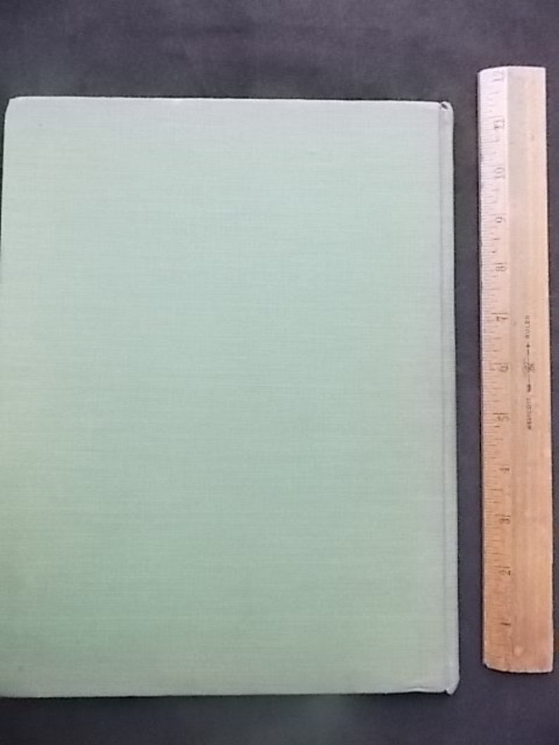 May include: A pale green book with a textured cover, next to a wooden ruler marked with inches. The book's cover is a solid, pale green colour. The ruler is made of wood and is marked with inch measurements.
