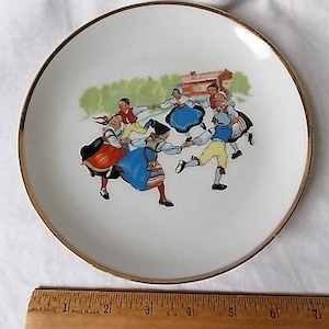 Peut inclure: Assiette en c&eacute;ramique blanche avec un bord dor&eacute;. L'assiette pr&eacute;sente une illustration color&eacute;e de personnes en costumes traditionnels dansant en cercle. L'assiette a une esth&eacute;tique vintage et mesure environ 18 cm de diam&egrave;tre.
