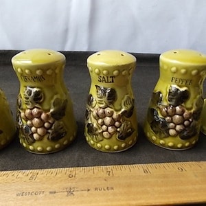 May include: A set of five ceramic salt and pepper shakers and a cinnamon shaker. The shakers are yellow with a grape design and the words "Salt", "Pepper", and "Cinnamon" printed on them. The shakers are approximately 3 inches tall.