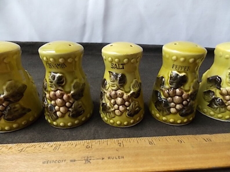 May include: A set of five ceramic salt and pepper shakers and a cinnamon shaker. The shakers are yellow with a grape design and the words "Salt", "Pepper", and "Cinnamon" printed on them. The shakers are approximately 3 inches tall.