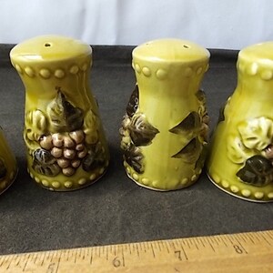 May include: A set of four ceramic salt and pepper shakers. The shakers are a light yellow-green color with a bulbous shape. Each shaker is decorated with a cluster of grapes and leaves in shades of brown and green.