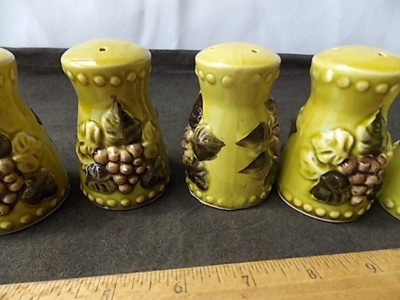 May include: A set of four ceramic salt and pepper shakers. The shakers are a light yellow-green color with a bulbous shape. Each shaker is decorated with a cluster of grapes and leaves in shades of brown and green.