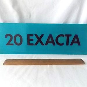 May include: A teal sign displaying the text "20 EXACTA" in dark purple. Beneath the sign is a wooden, rectangular object. The background is a white, creased fabric.