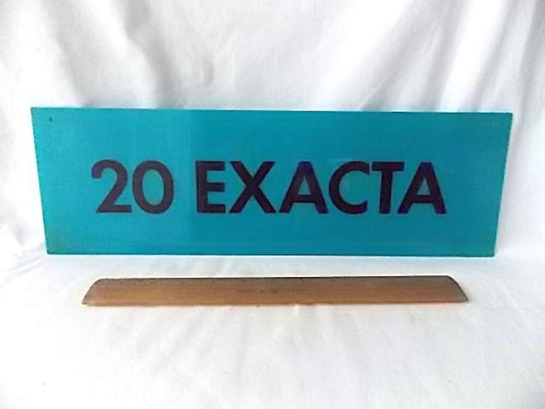 May include: A teal sign displaying the text "20 EXACTA" in dark purple. Beneath the sign is a wooden, rectangular object. The background is a white, creased fabric.