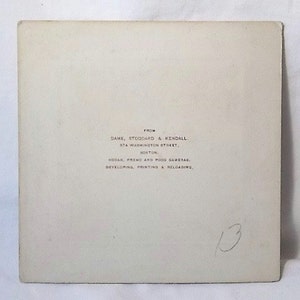 K&ouml;nnte beinhalten: Eine quadratische, cremefarbene Papierkarte mit schwarzem Aufdruck: FROM DAME, STODDARD & KENDALL, 374 WASHINGTON STREET, BOSTON, KODAK, PREMO AND POCO CAMERAS, DEVELOPING, PRINTING & RELOADING. Die Zahl 13 ist handschriftlich mit Bleistift notiert.