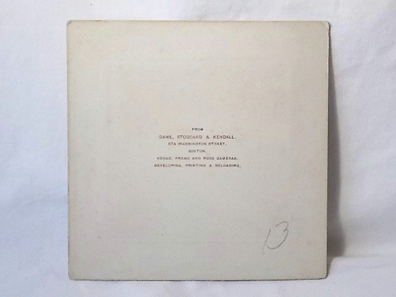 K&ouml;nnte beinhalten: Eine quadratische, cremefarbene Papierkarte mit schwarzem Aufdruck: FROM DAME, STODDARD & KENDALL, 374 WASHINGTON STREET, BOSTON, KODAK, PREMO AND POCO CAMERAS, DEVELOPING, PRINTING & RELOADING. Die Zahl 13 ist handschriftlich mit Bleistift notiert.