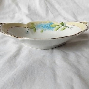 May include: A small, white porcelain dish with a scalloped edge and two small handles. The rim is trimmed with gold. The interior features a hand-painted design of blue flowers and green leaves against a pale yellow background.