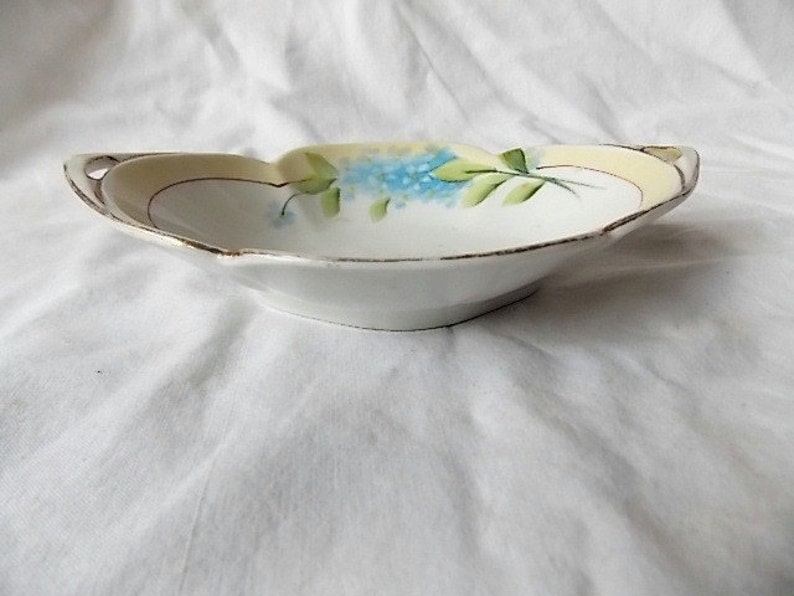 May include: A small, white porcelain dish with a scalloped edge and two small handles. The rim is trimmed with gold. The interior features a hand-painted design of blue flowers and green leaves against a pale yellow background.