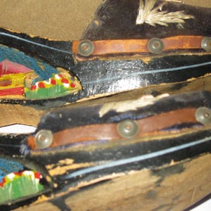 Peut inclure: Une paire de sandales &agrave; plateforme en bois noir avec une bride en cuir marron fix&eacute;e par des clous m&eacute;talliques. Le c&ocirc;t&eacute; de la plateforme est orn&eacute; d'un paysage peint &agrave; la main en bleu, jaune, rouge et vert. La bride sup&eacute;rieure pr&eacute;sente des coutures blanches.