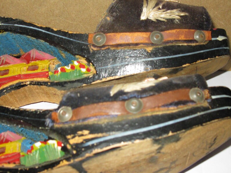 Peut inclure: Une paire de sandales &agrave; plateforme en bois noir avec une bride en cuir marron fix&eacute;e par des clous m&eacute;talliques. Le c&ocirc;t&eacute; de la plateforme est orn&eacute; d'un paysage peint &agrave; la main en bleu, jaune, rouge et vert. La bride sup&eacute;rieure pr&eacute;sente des coutures blanches.