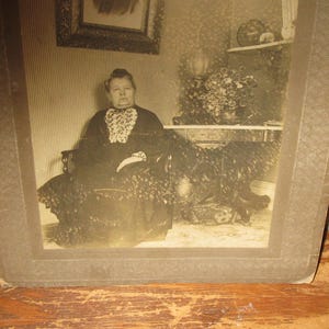 May include: Sepia-toned photograph of a woman seated in a decorative chair. She wears a dark dress with a white lace collar. The photograph is framed in a brown, patterned border. A painting and a shelf with decor are visible in the background.