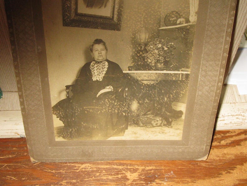May include: Sepia-toned photograph of a woman seated in a decorative chair. She wears a dark dress with a white lace collar. The photograph is framed in a brown, patterned border. A painting and a shelf with decor are visible in the background.